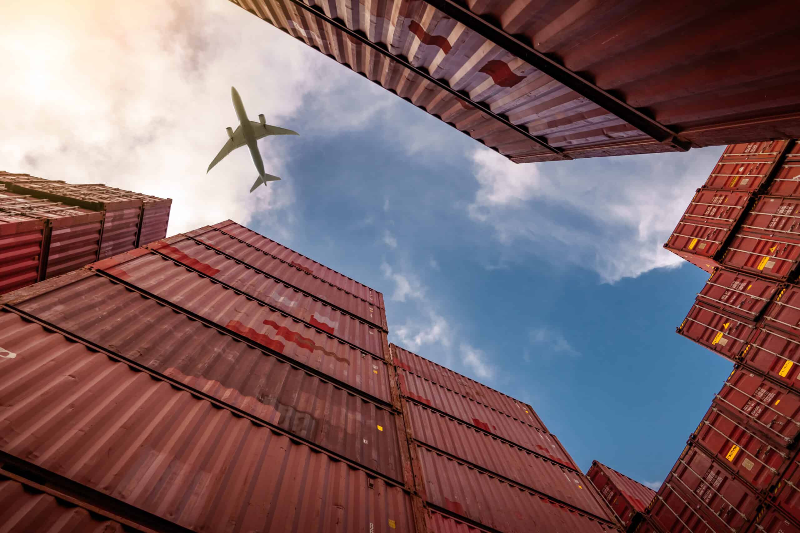 A low-angle view of stacked shipping containers with a cargo plane flying overhead, symbolizing global logistics and the supply chain planning process.