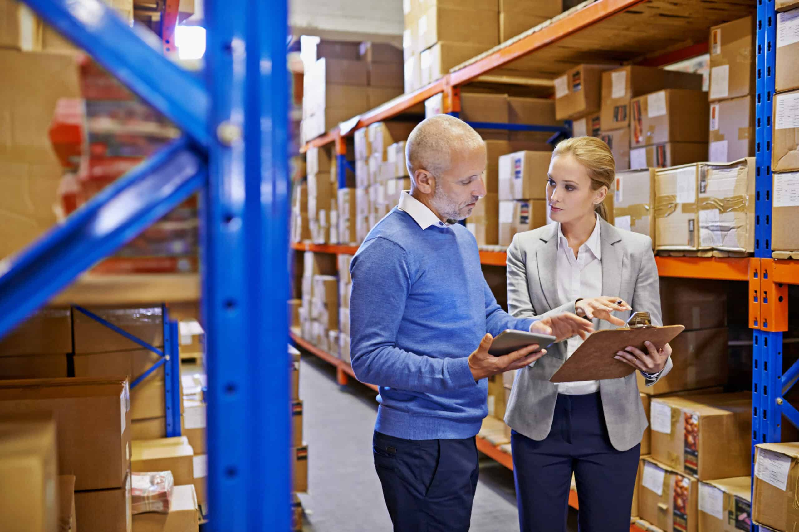 Two supply chain professionals reviewing inventory plans in a warehouse, discussing data on a tablet and clipboard with shelves of boxes in the background.