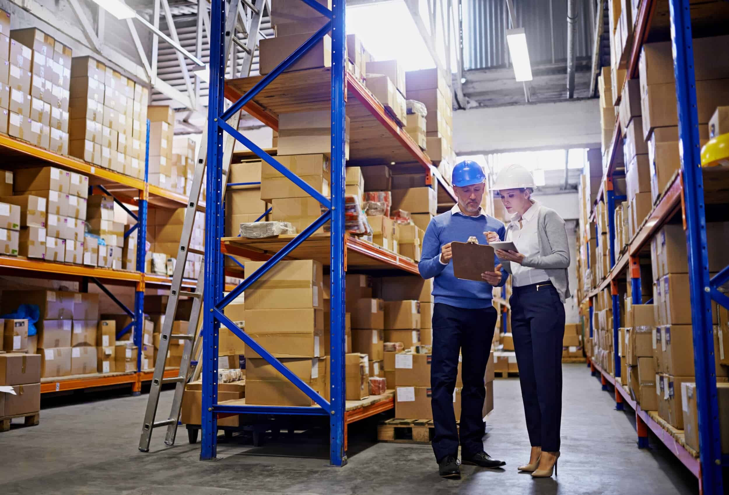 Two workers wearing safety helmets reviewing inventory on a clipboard inside a warehouse filled with stacked boxes on shelves.