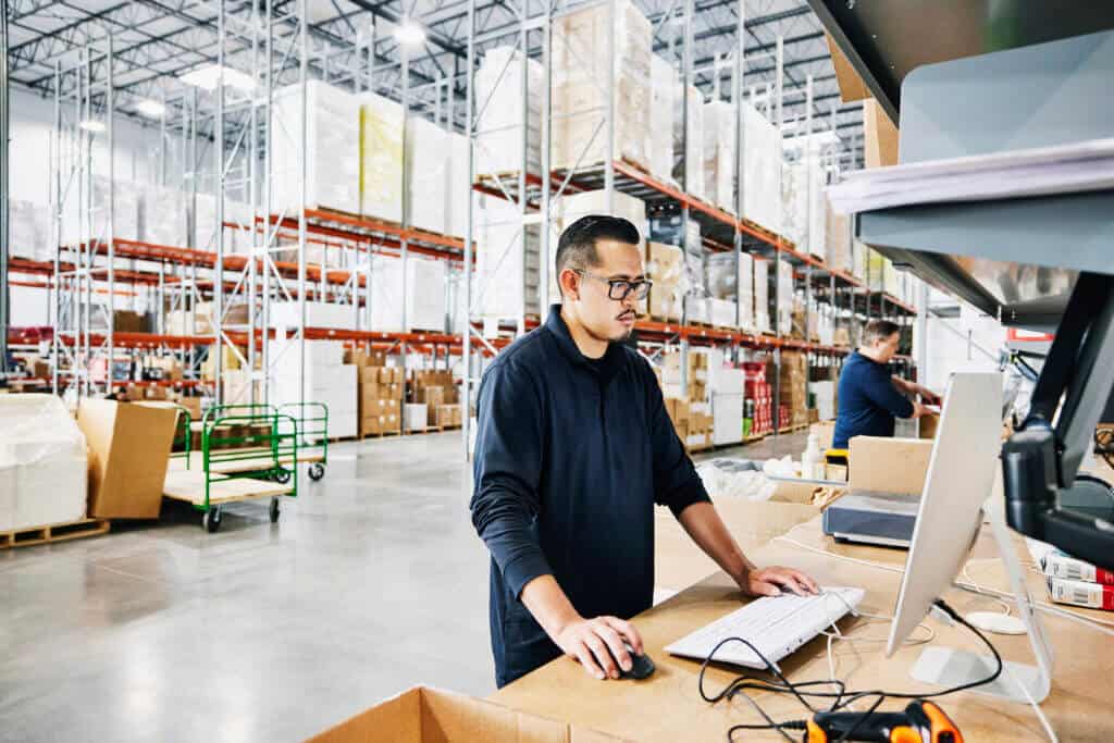 Medium wide shot of male warehouse worker checking orders at computer workstation in warehouse Medium wide shot of male warehouse worker checking orders at computer workstation in warehouse