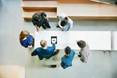 An overhead view of a team of people standing at a table looking at a tablet