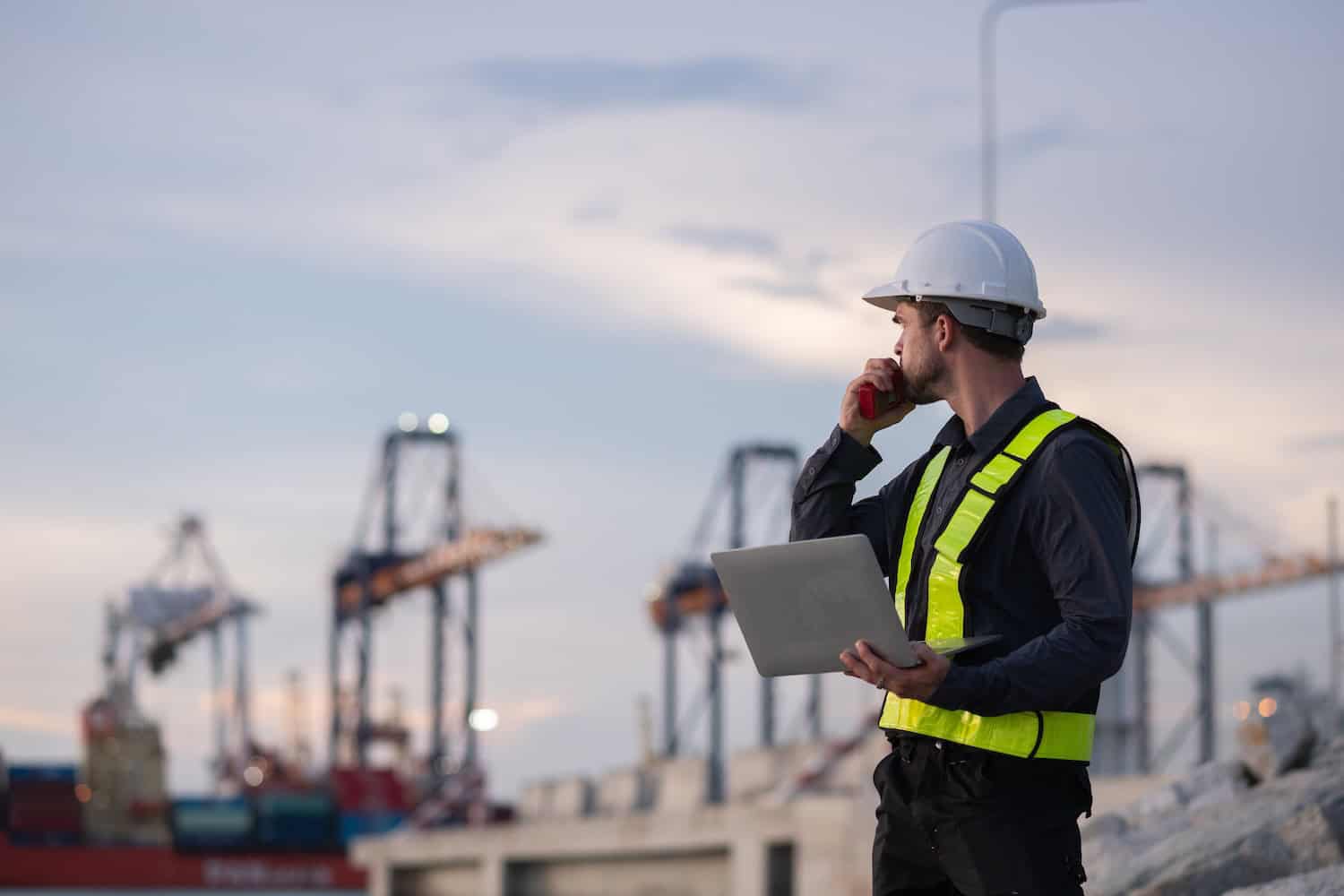 A supply chain manager wearing a safety vest and hard hat stands at a shipping port with cranes in the background, holding a laptop and talking on a radio while overseeing operations and planning activities