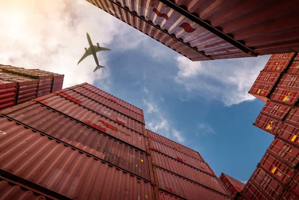 A low-angle view of stacked shipping containers with a cargo plane flying overhead, symbolizing global logistics and the supply chain planning process.
